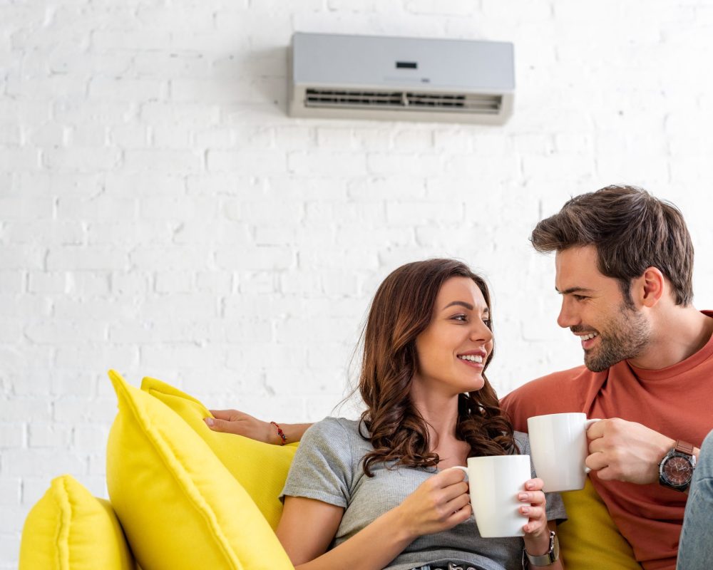 happy couple with cups sitting on sofa under air conditioner at home
